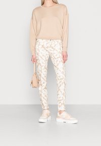 Sudadera beis, pantalones blancos con patrones abstractos en marrón claro y zapatillas beis. El modelo sostiene un pequeño bolso beis.