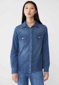 Camicia in denim di blu medio con maniche lunghe, chiusura con bottoni sul davanti e due tasche sul petto con dettagli hardware decorativi.