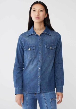 Camicia in denim di blu medio con maniche lunghe, chiusura con bottoni sul davanti e due tasche sul petto con dettagli hardware decorativi.