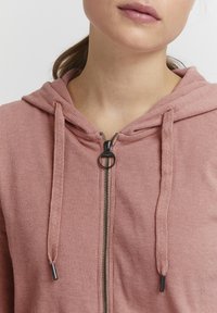 Svetloružová rypsová mikina na zips s kapucňou na sťahovanie a čiernym zipsom. Má voľný strih a štruktúrovanú tkaninu.