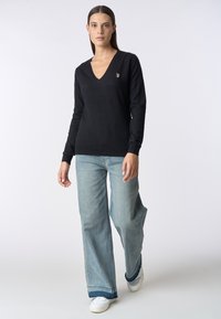 Maglione nero a v-neck realizzato in tessuto morbido, con maniche lunghe e un piccolo logo come accentuazione. Abbinato a jeans a gamba larga blu chiaro e scarpe bianche.