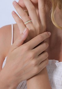 Zlatý prsteň s viacerými prúžkami, zdobený iridiscenčnými kameňmi a akcentmi v tvare krútenej šnúrky. Ruky sú zložené dohromady, s viditeľným svetlým tónom pleti.
