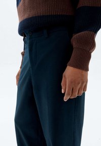 Eine Person trägt maßgeschneiderte marineblaue Hosen und einen braun-blauen, gerippten Strickpullover und steht mit entspannter Haltung.