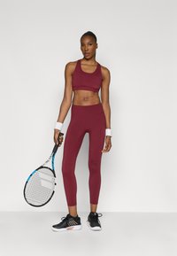 Bordó sportmelltartó és hozzáillő, magas derekú leggings rugalmas anyagból, fekete sportcipővel és teniszekével a kezében.