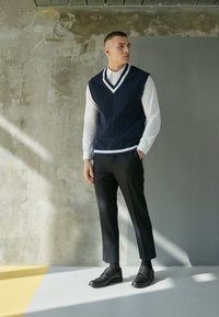 Veste sans manches en maille torsadée marine portée sur une chemise blanche, associée à un pantalon noir sur mesure et des chaussures en cuir noir, présentant un col en V.