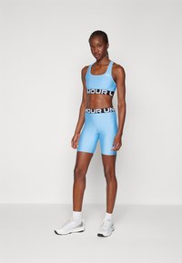 Svijetloplava atletska sportska grudnjak i visoki šorc s crnim pojasom na kojem stoji "UNDER ARMOUR" u bijeloj boji. Cipele su bijele s sivoj detaljima.