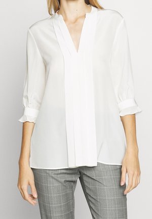 Mujer con blusa blanca de mangas 3/4 y pantalones a cuadros grises, mostrada desde el cuello hasta la mitad del muslo sobre un fondo liso.