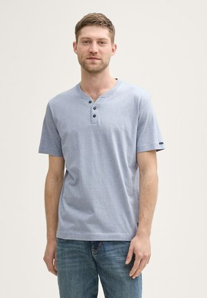 Young man with short hair wearing a light blue short-sleeve henley shirt and blue jeans, standing against a plain light background.