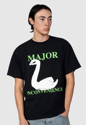 Młody mężczyzna o średniej długości ciemnych włosach nosi czarną koszulkę z białym łabędziem i zielonym napisem: "WIĘKSZA NIEWYGODA."