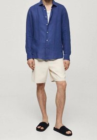 Chemise à boutons en lin bleu, t-shirt blanc en dessous, short beige et sandales noires à enfiler. Design simple avec une coupe décontractée.