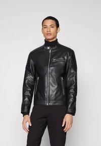 Black leather jacket with a zip closure, stand collar, ribbed shoulder accents, and two side zip pockets, worn over a black turtleneck.