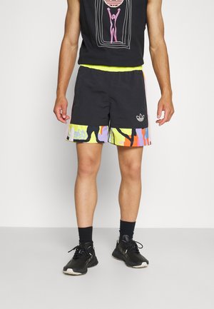 Hombre con pantalones cortos deportivos negros con patrones abstractos coloridos en el dobladillo, zapatillas negras, calcetines negros y una camiseta sin mangas negra con un diseño gráfico.