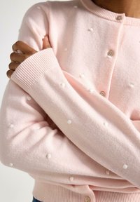 Cardigan rosa chiaro con pois bianchi in rilievo, chiusura con bottoni e polsini a coste. Materiale morbido, vestibilità rilassata e design semplice.