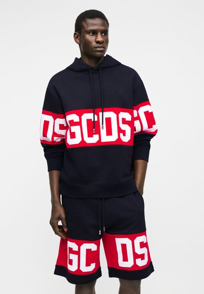 Άνδρας που φοράει μαύρο φούτερ με κουκούλα και σορτς με έντονες κόκκινες και λευκές λωρίδες λογότυπου GCDS στο στήθος, στα μανίκια και στα πόδια, στέκεται μπροστά σε μονόχρωμο φόντο.