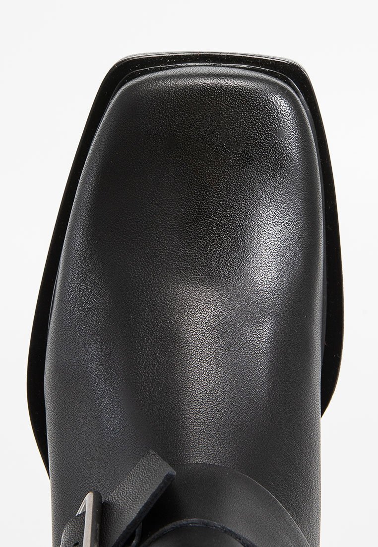 Bottine en cuir noir avec un bout carré, texture lisse et accent de boucle sur le côté. Présente un petit talon et un design minimal.