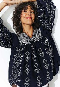 Blouse brodée bleu marine avec des motifs floraux et géométriques blancs complexes, présentant un décolleté en V et des manches bouffantes.