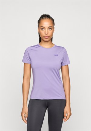 Kobieta stoi twarzą do przodu, ubrana w fioletowy t-shirt z krótkim rękawem i czarne legginsy na jednolitym jasnym tle.