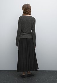 Femme aux longs cheveux se tient face à un mur gris, portant un pull en maille gris foncé, une jupe plissée noire et des chaussures plates noires.