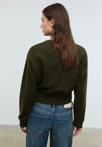 Donna con lunghi capelli castani indossa un maglione verde scuro e jeans blu, in piedi con le spalle rivolte verso uno sfondo grigio chiaro uniforme.