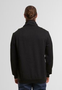 Mann mit braunen Haaren im Dutt, der eine schwarze strukturierte Jacke und dunkelblaue Hosen trägt und mit dem Rücken zu einem schlichten Hintergrund steht.