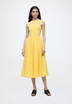 Kobieta o długich ciemnych włosach, ubrana w żółtą sukienkę midi bez rękawów i czarne mule na szerokim obcasie z odkrytymi palcami, stoi na białym tle.