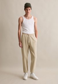 Beige joggerbroeken met elastische boorden, trekkoord in de taille, zijzak; gecombineerd met een witte tanktop en een groene gebreide beanie, staand op een neutrale achtergrond.