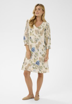 Kvinde iført en knælængde blomsterkjole med lange ærmer og beige flettede slingback-flade sko, stående mod en hvid baggrund.