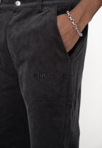 Pantaloni din catifea neagră cu rebie verticală, având buzunare laterale și un logo vizibil al brandului imprimat pe țesătură. Brățară argintie la încheietură.