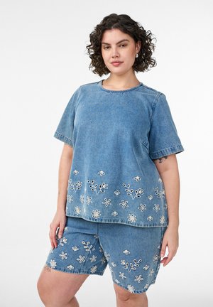 Kvinde med krøllet hår iført blå denim top med korte ærmer og matchende shorts, begge broderet med hvide blomster ved kanterne.