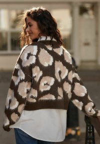 Sweter w brązowo-kremowy wzór w panterkę, o luźnym kroju, szerokich rękawach i prążkowanym dolnym obszyciu, nałożony na białą koszulę. Teksturowana wełniana tkanina.