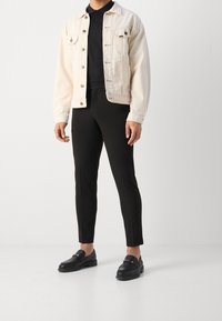 Light beige denim jacket, black polo shirt, tailored black pants, and black loafers with gold hardware accents.