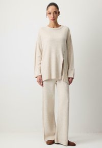 Pulover tricotat bej cu textură în relief, cu fante laterale și două butoane la margine. Asortat cu pantaloni largi din tricot cu textură în relief.