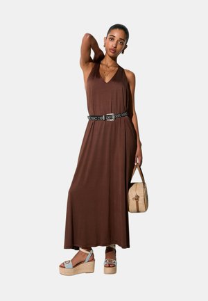 Donna con abito lungo marrone senza maniche con cintura nera borchiata, che tiene una borsa intrecciata e indossa sandali con zeppa, in piedi con il braccio alzato.