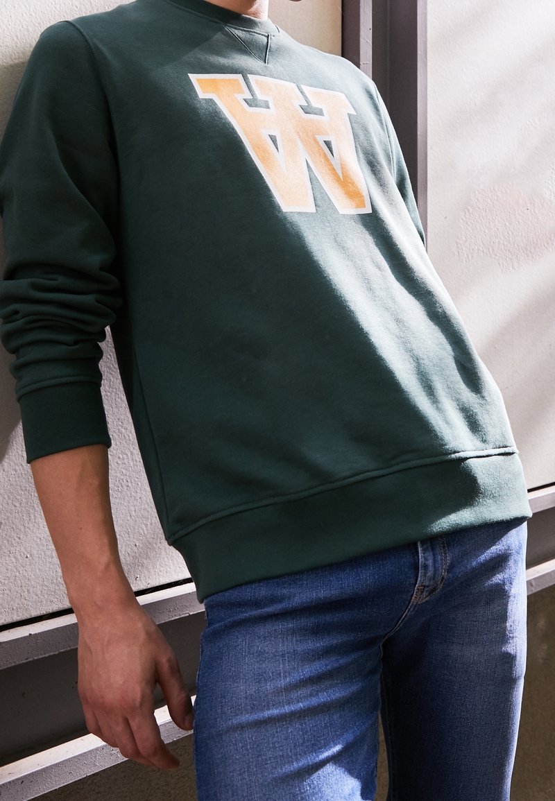 Zelená mikina s bílým a oranžovým logem "W", s kulatým výstřihem a žebrovanými manžetami, kombinovaná s modrými džíny.