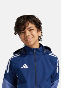 Giacca atletica blu navy con strisce bianche sulle maniche, chiusura con zip e cappuccio. Texture liscia, realizzata in materiale leggero.