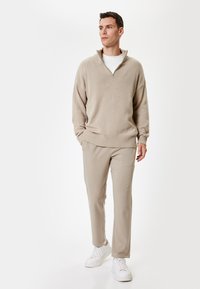 Beige strikket sweater med halvlange lynlås, parret med lyse beige bukser og hvide sneakers. Enkel, afslappet pasform med ribbede detaljer.