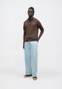 Brūns adīts džemperis ar rāvējslēdzēja apkakli, saskaņots ar gaiši ziliem plaša stila džinsiem un zaļiem sporta apaviem. Minimālistisks dizains, bez redzamām rakstām.