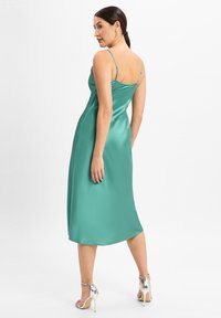 Robe midi en satin vert avec de fines bretelles ajustables, une silhouette épurée et une texture lisse. Portée avec des sandales à talons argentées.