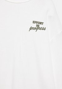 Tissu tricoté blanc avec un texte vert foncé sur la poitrine indiquant « EFFORT IN progress » en polices gras et cursives.
