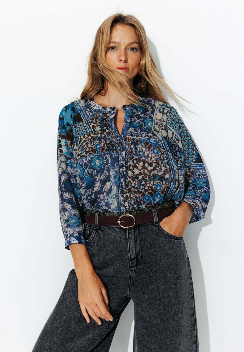 Blouse légère multicolore avec des motifs floraux et géométriques complexes, à encolure boutonnée, avec des manches larges, associée à un jean noir taille haute.