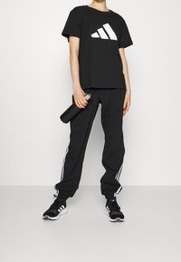 Czarny t-shirt z dużym białym logo Adidas, w połączeniu z czarnymi spodniami z białymi paskami po bokach oraz czarnymi butami sportowymi. Trzyma butelkę wody.