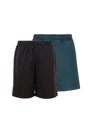 Homme portant un short noir jusqu'aux genoux, une chemise blanche et des chaussures de sport argentées, debout les mains dans les poches sur fond gris.