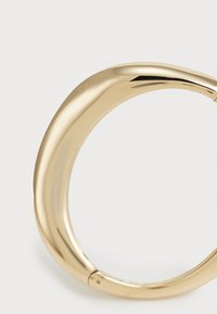 Aukso tono bangles apyrankė su lygiu, kreivu dizainu ir subtiliai išgraviruotu logotipu. Paviršius poliruotas, elegantiškai atspindintis šviesą.