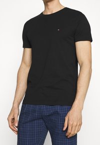 Camiseta de algodón negra con cuello redondo y mangas cortas. Pequeño logo con acentos rojos y blancos en el lado izquierdo del pecho. Diseño sencillo.