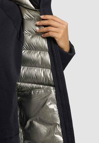 Marineblauwe buitenjas met een glanzende zilveren geïsoleerde voering, voorzien van doorgestikte delen en een gladde textuur, gedeeltelijk open om het interieur te tonen.