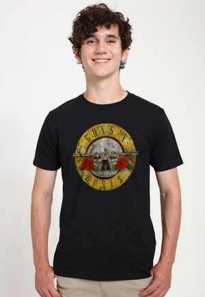 Young man with curly dark hair wearing a black Guns N' Roses T-shirt and beige pants, smiling against a plain background.