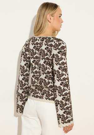 Donna bionda che indossa un maglione crema con motivi floreali marroni e neri, abbinato a pantaloni bianchi, in piedi davanti a uno sfondo bianco.