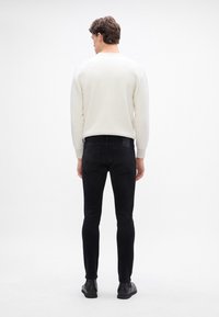 Jersey blanco de canalé con cuello redondo, combinado con jeans negros ajustados. El modelo lleva botas negras hasta el tobillo y está de espaldas a la cámara.