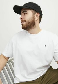 Vit kortärmad T-shirt med en svart "&"-logotyp på vänster bröst, i kombination med olivgröna shorts och en svart keps. Slät textur.