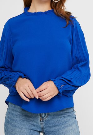 Vrouw met een felblauwe blouse met geplooide lange mouwen en gerimpelde manchetten, gecombineerd met lichtblauwe jeans, handen voor zich geklemd.